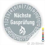 Prüfplaketten rund, Farbe- Grau, Text "Nächste Gasprüfung" in Weiß danach eine Flamme als Pictogramm oder Icon in Weiß, Startjahr- 24, Monatszahlen Oben und Jahreszahlen Unten in Kreisform.