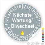 Prüfetiketten, Prüfaukleber rund, Farbe- Grau, Text "Nächste Wartung/Ölwechsel" in Weiß. Startjahr- 24, Monatszahlen Oben und Jahreszahlen Unten in Kreisform, über den Jahreszahlen ein Öltropfen als Icon/Piktogramm in Gelb.