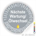 Prüfetiketten, Prüfaukleber rund, Farbe- Grau, Text "Nächste Wartung/Ölwechsel" in Weiß. Startjahr- 26, Monatszahlen Oben und Jahreszahlen Unten in Kreisform, über den Jahreszahlen ein Öltropfen als Icon/Piktogramm in Gelb.