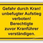 Warnschild vor Kran mit der Aufschrift "Gefahr durch Kran! Unbefugter Aufstieg verboten! Berechtigte zuvor Kranführer verständigen." in gelber Farbe.