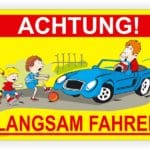 Illustration eines Verkehrsschildes mit der Aufschrift "Achtung! Langsam fahren". Auf dem Schild sind Kinder zu sehen, die mit einem Ball spielen, während ein Fahrer in einem blauen Auto langsam vorbeifährt.