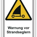 Warnschild mit der Aufschrift "Warnung vor Strandseglers" auf gelbem Hintergrund, das auf die Anwesenheit von Strandseglern hinweist und Sicherheitshinweise für Fußgänger gibt.