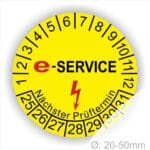 Prüfplaketten rund, Farbe- Gelb, Text "E-SERVICE" in Schwarz danach ein Elektro- Pfeil in Rot, Startjahr- 25, Monatszahlen Oben und Jahreszahlen Unten in Kreisform. Über den Jahreszahlen Text "Nächster Prüftermin"