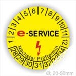 Prüfplaketten rund, Farbe- Gelb, Text "E-SERVICE" in Schwarz danach ein Elektro- Pfeil in Rot, Startjahr- 28, Monatszahlen Oben und Jahreszahlen Unten in Kreisform. Über den Jahreszahlen Text "Nächster Prüftermin"