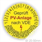 Prüfplaketten rund, Farbe- Gelb, Text "Geprüft PV-Anlage nach VDE" in Schwarz danach ein Elektro- Pfeil in Rot, Startjahr- 26, Monatszahlen Oben und Jahreszahlen Unten in Kreisform. Über den Jahreszahlen Text "Nächster Prüftermin"