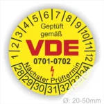 Gelbes Prüfzeichen mit der Aufschrift "Geprüft gemäß VDE" und den Nummern 0701-0702, umgeben von einer Skala für den nächsten Prüftermin. Durchmesser von 20-50 mm.