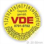 Gelbes VDE Prüfplakette mit der Aufschrift "Geprüft gemäß VDE" und der Prüfnorm 0701-0702. Die Plakette zeigt einen nächsten Prüftermin und einen Durchmesser von 20-50 mm.