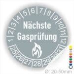 Prüfplaketten rund, Farbe- Grau, Text "Nächste Gasprüfung" in Weiß danach eine Flamme als Pictogramm oder Icon in Weiß, Startjahr- 25, Monatszahlen Oben und Jahreszahlen Unten in Kreisform.