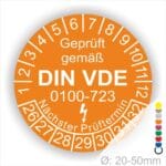 Prüfetiketten rund, Farbe- Orange, Text "Geprüft gemäß VDE 0100-0723" in Weiß danach ein Elektro- Pfeil in Weiß, Startjahr- 26, Monatszahlen Oben und Jahreszahlen Unten in Kreisform. Über den Jahreszahlen Text "Nächster Prüftermin"
