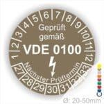 Prüfaukleber rund Farbe- Braun, Text "Geprüft gemäß VDE 0100" in Weiß danach ein Elektro- Pfeil in Weiß, Startjahr- 27, Monatszahlen Oben und Jahreszahlen Unten in Kreisform. Über den Jahreszahlen Text "Nächster Prüftermin"