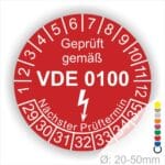 Prüfetikette rund, Farbe- Rot, Text "Geprüft gemäß VDE 0100" in Weiß danach ein Elektro- Pfeil in Weiß, Startjahr- 29, Monatszahlen Oben und Jahreszahlen Unten in Kreisform. Über den Jahreszahlen Text "Nächster Prüftermin"