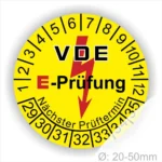 VDE E-Prüfungsplakette mit gelbem Hintergrund, die den nächsten Prüftermin anzeigt. Die Plakette hat einen Durchmesser von 20-50 mm und zeigt die Prüfzahlen von 1 bis 35. Ein Blitzsymbol ist in der Mitte abgebildet, das auf elektrische Sicherheit hinweist.