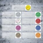 Runde DGUV-V3-Kabelprüfplaketten in verschiedenen Farben und Startjahren auf grauem Hintergrund, jeweils mit Monatssegmenten, Prüfvermerk ‚Geprüft‘ und Blitzsymbol zur Kennzeichnung der gesetzlichen Elektrogeräteprüfung.