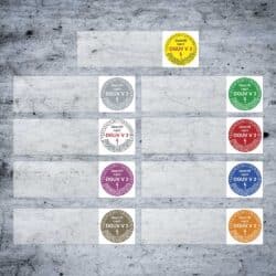 Runde DGUV-V3-Kabelprüfplaketten in verschiedenen Farben und Startjahren auf grauem Hintergrund, jeweils mit Monatssegmenten, Prüfvermerk ‚Geprüft‘ und Blitzsymbol zur Kennzeichnung der gesetzlichen Elektrogeräteprüfung.