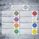 Prüfplaketten mit farbigen Texten. Diese Prüfplaketten für Kabel sind in verschiedenen Farben dargestellt: Weiß, Bronze, Rot, Lila, Orange, Grün und Gelb. Jede Plakette enthält Text, der das Jahr und den Vermerk "Geprüft nach DGUV V 3" zeigt.